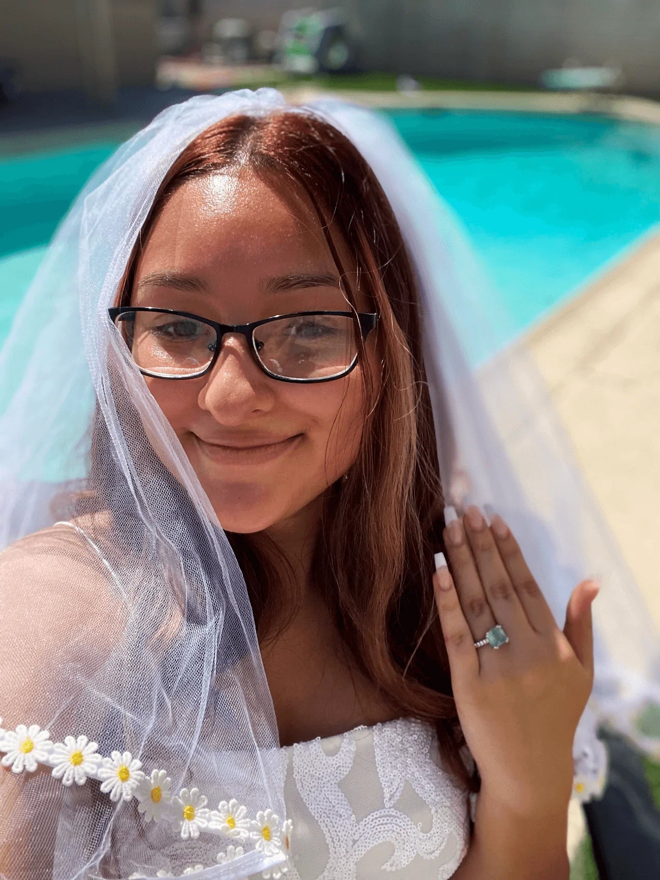 Woman in a wedding dress with a veil and flowers, standing by a pool.