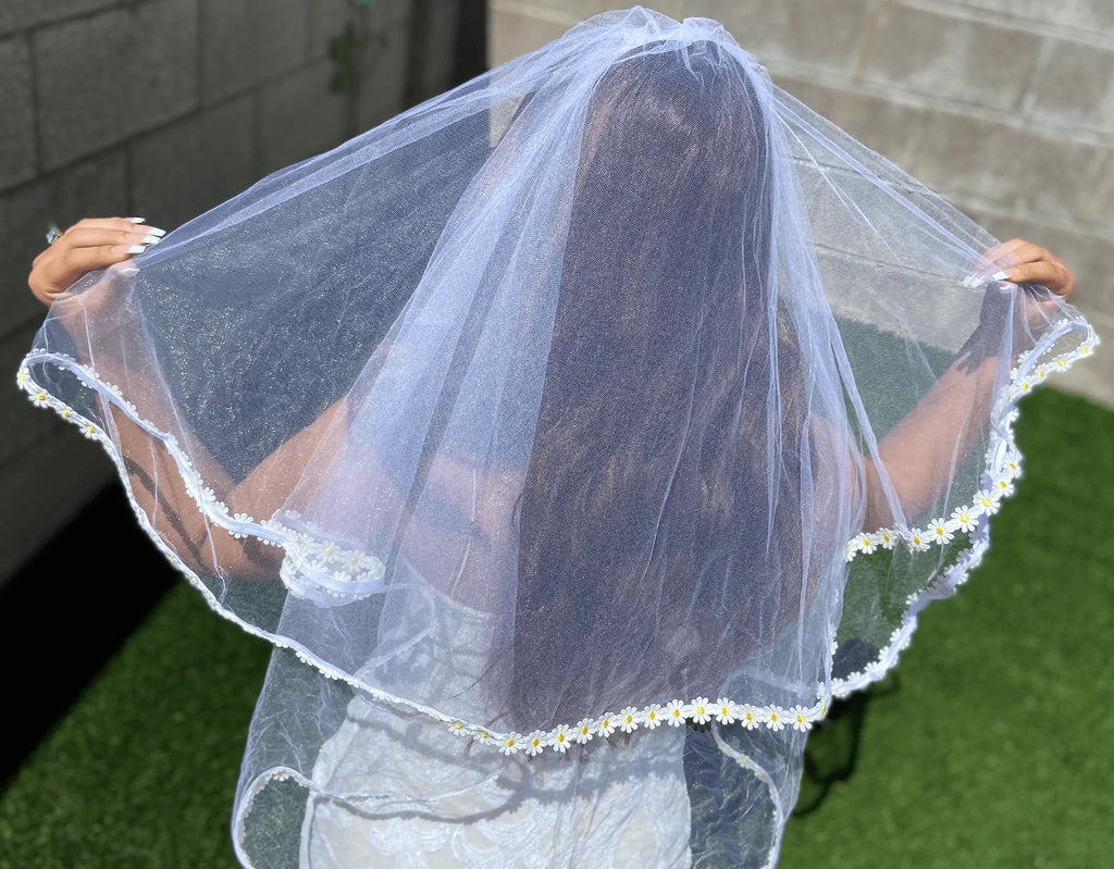 Person holding a white veil with floral trim outdoors.